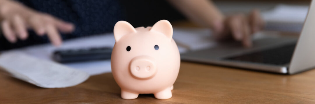 Piggybank on table of business woman calculating money, doing accounting, paperwork job, using laptop computer, calculator in blurred background. Investment, savings, currency concept. Banner - Powered by Adobe