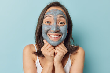 Portrait of cheerful Asian woman applies clay facial mask to refine pores and clear skin smiles toothily keeps hands under chin wears t shirt isolated over blue background wants to be always beautiful