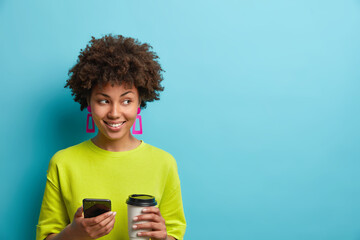 Positive pleasant looking woman holds smartphone and coffee to go scrolls newsfeed and enjoys favorite drink wears casual t shirt focused away isolated over blue background empty space for your text