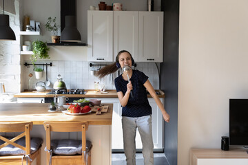 Joyful happy dancer girl enjoying cooking in kitchen, dancing to music from wireless headphones, singing sings at ladle, laughing, having fun at table with fresh vegetables