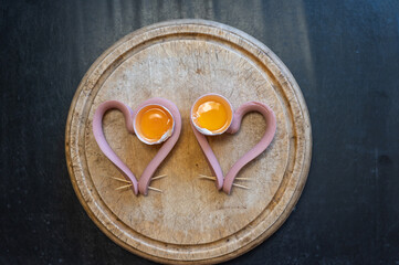 Two heart-shaped sausages lie on a cutting board. Raw chicken yolks in egg shells lie side by side....