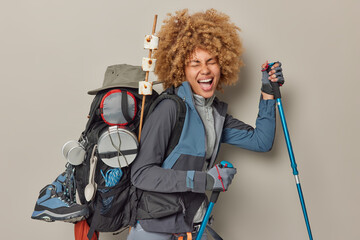 Happy curly haired female camper enjoys hiking trip holds trekking poles tries to reach top of...
