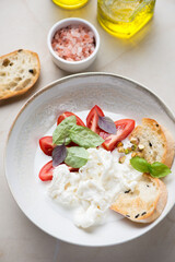 Plate with stracciatella or stretched curd fresh cheese served with ciabatta, red tomatoes and basil leaves, vertical shot on a beige stone background