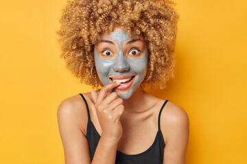 Positive surprised European woman uses new beauty product applies clay mask on face to cleanse smooth and brighten skin bites finger wears black t shirt isolated over vivid yellow background