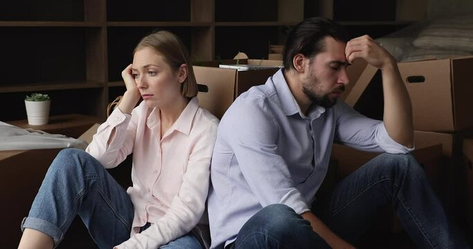 Tired sad homeowner man and woman exhausted with relocation, packing, unpacking cardboard boxes, sitting on floor of new home at paper containers. Couple moving out due to divorce, breakup