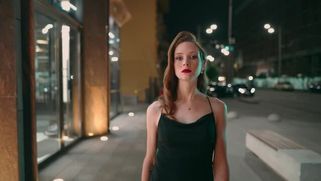 Stylish beautiful young woman in a black dress walks through the night city and looks at the camera