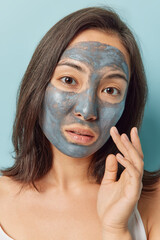 Close up shot of serious brunette young woman with eastern appearance touches skin applies clay mask to nourish skin and eliminate wrinkles looks directly at camera poses against blue background