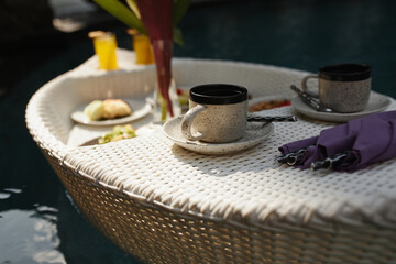 Cup of coffee on a floating tray in the pool. Floating breakfast in the swimming pool. Straw tray with different food.