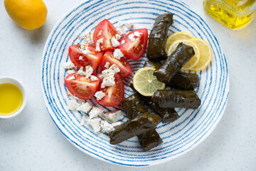Greek dolma or dolmades with red tomatoes, feta and lemon on a blue and white plate, elevated view on a white stone background