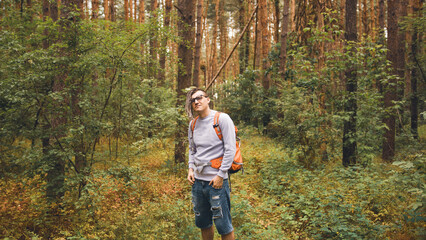 Obraz premium A man with a backpack poses in the woods. A male traveler with dreadlocks standing in a summer forest with a backpack