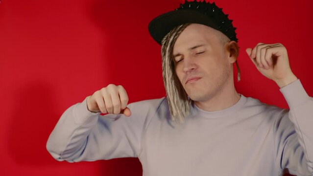 Young handsome man with dreadlocks in black cap dancing on red background. Relaxed male enjoying music, closing eyes, dancing in studio.

