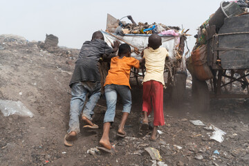 Three black street boys pushing a heavy garbage cart up a heap in urban landfill; informal waste...