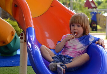 Fototapeta na wymiar Handsome boy eats candy on a stick. Children's sweets. Playground. Colorful slide