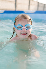 Naklejka premium Little baby in swimming goggles, girl swim in paddling pool. Diving kid. Learning child to swim. Summer leisure and family holidays and vacation concept. People swim in a metal frame pool in backyard.