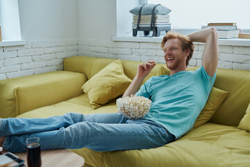Obraz premium Happy young redhead man enjoying snacks while sitting on the couch at home