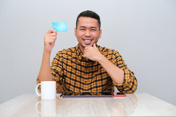 Adult Asian man sitting in front of mobile gadget with happy expression while showing blank credit card