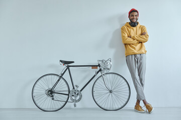Naklejka premium Happy young African man leaning at the wall while standing near his bicycle
