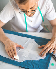 A self-taught seamster in white t-shirt and glasses works with fabric, pattern and chalk. A tailor transfers a template to fabric in a home studio. Hobby, small business and life style concept