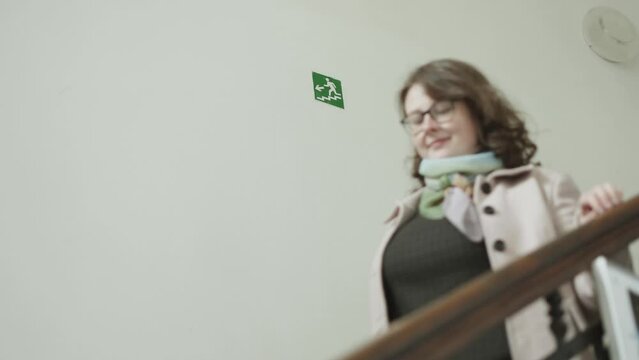 Green evacuation sign with arrow, man and staircase indicating the exit direction on a white wall in a building. A young woman in glasses goes down the stairs, slow motion.