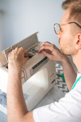 Obraz premium A blond man with glasses changes a bobbin in a sewing machine. A tailor with a measuring tape on his neck in a white t-shirt. The concept of small business, lifestyle and enjoying of work