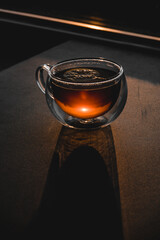 Close up of tea with lemon in transparent cup and sun glare on table. Hot drink of sparkling orange liquid in double glass cup with hard shadows and sunlight reflection, shine in line of sun light.