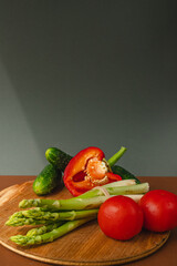 Vegetables lie on a wooden board: tomatoes, asparagus, cucumbers, red bell peppers. brown, dark gray background. place for text.