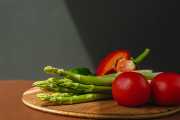 Vegetables lie on a wooden board: tomatoes, asparagus, cucumbers, red bell peppers. brown, dark gray background. place for text.
