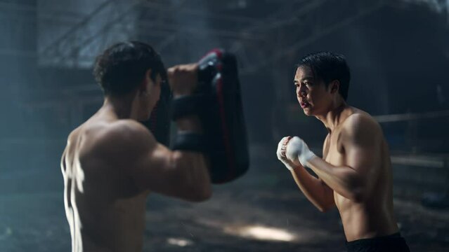 4K Strong Asian sportsman practicing boxing workout punching with male combat sport trainer in abandoned building. Active man coach support male boxer do fighting exercise kickboxing in dark old gym