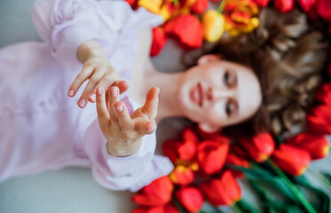 Obraz na płótnie Canvas A young woman is lying on the floor among red tulips. The concept of March 8, Valentine's Day. Spring portrait of a woman.Focus on the girl's hands, in the foreground.