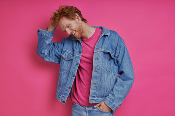 Handsome redhead man in denim clothing holding hand ion hair against pink background