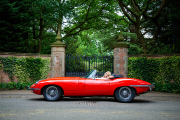 Lady with long blond hair resting in a red E-Type Jaguar with gates and park in the background - Powered by Adobe