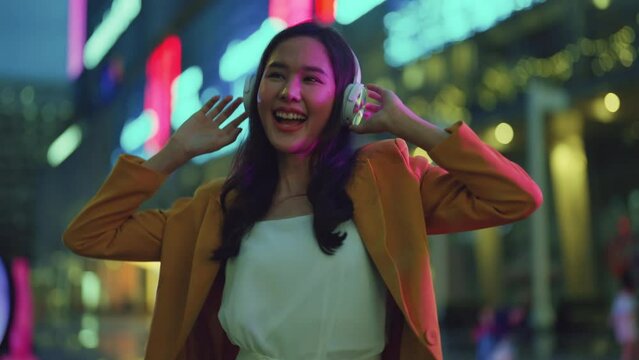 Trendy dressed asian young woman feeling joyful carefree dancing listening music song in earphones, happy woman dancing freedom move having fun on evening city street with neon illumination building