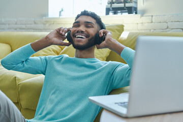 Joyful African man in headphones enjoying music while sitting on the floor at home