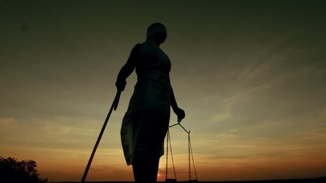 Woman in costume of goddess justice holds sword and scales, she raises scales against the background of sunset, bottom view. Concept justice