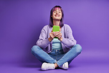 Beautiful young woman holding smart phone and smiling while sitting against purple background