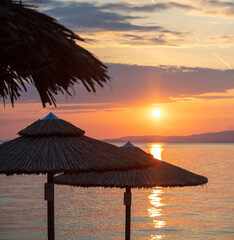 Fototapeta premium Sunset on the beach. Greece. Straw umbrella silhouette, golden reflection on rippled ocean water.