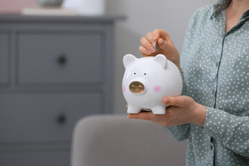 Young woman putting coin into piggy bank indoors, closeup