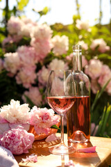 Bottle and glass of rose wine near beautiful peonies on wooden table in garden