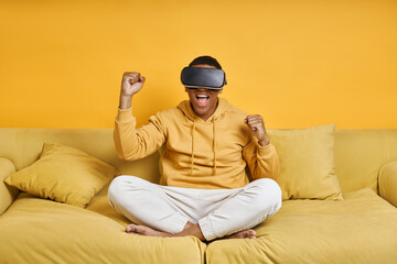 Happy young man in virtual reality glasses gesturing while sitting against yellow background