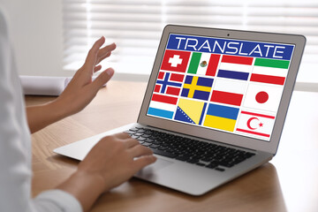 Translator using modern laptop with images of different flags on screen at wooden table indoors,...