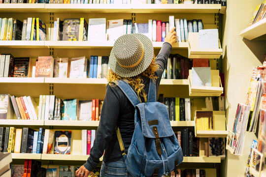 Back view of woman with hat and backpack choosing a book to buy inside a library or newspaper store. People prepare to travel. Leisure and shopping female people activity. Traveler lifestyle