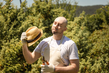 Sunstroke. Portrait of bald bearded man wearing in gloves and apron fanning with straw hat holding...