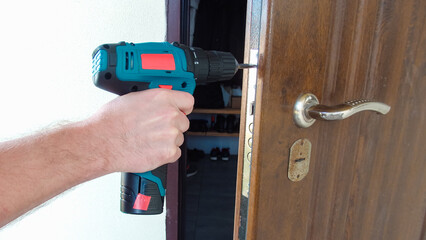 Hand 's man with screwdriver. The worker installs a door lock in the front door, metal doors with a polymer coating using an electric drill screwdriver, close-up.