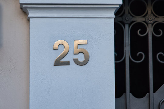 The number '25' is mounted in big golden letters on a white column next to a steel gate.  Luxurious house exterior. 