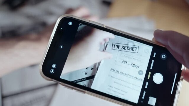 Spy holds mobile phone photographing sensitive classified documents on a desk. Man or spy or mole using smartphone to take pictures of leaked top secret government papers or military operation memos