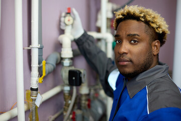 Confident African-American who loves his job checks the pressure on the sensor in the boiler room. Black man in overalls professionally maintains the boiler room equipment.