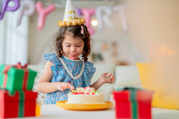 Birthday. Joyful, anniversary dressed kid daughter in party hat enjoys finger tasty sweet cake for...