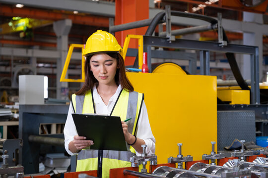 Selective focus at young women inspector wearing safety equipment. While doing audit for quality and safety control inside of factory area. With blurred background of heavy machine.