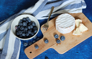 Brie cheese on wooden boars, small fork, bowl of blueberry, striped tea towel on textured background with copy space. Eating fresh concept. 