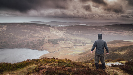 Man from behind looking towards Lough Tay (Guinness Lake) from Luggala peak in the Wicklow mountains Ireland. Cloudy and wet day